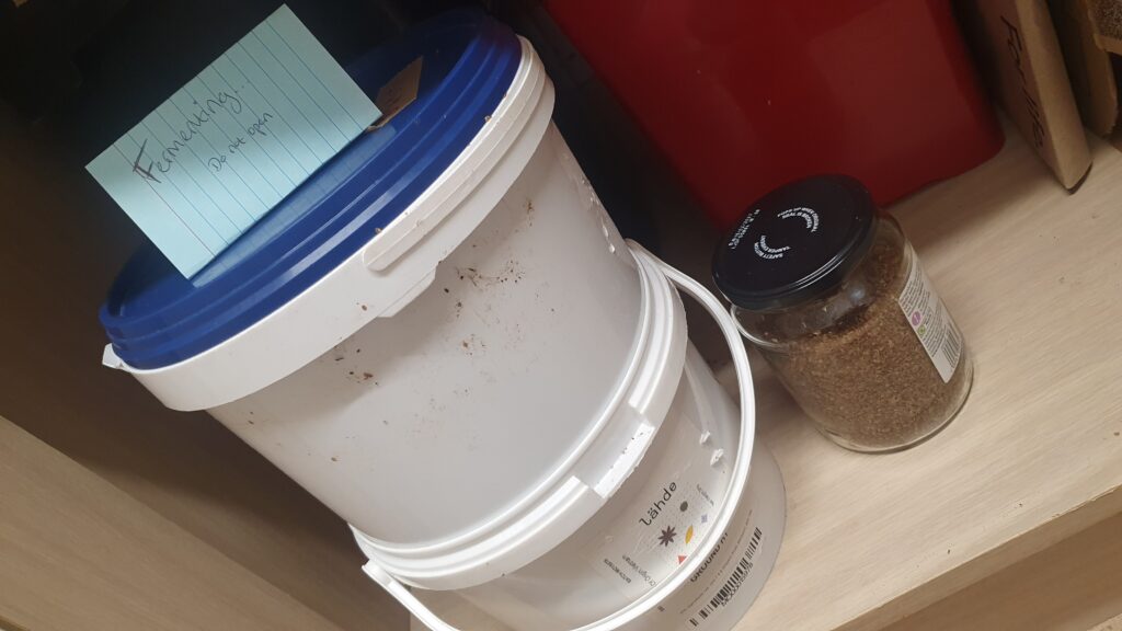 Two white buckets stacked on top of each other, with a blue lid, and a sign saying 'fermenting do not open'