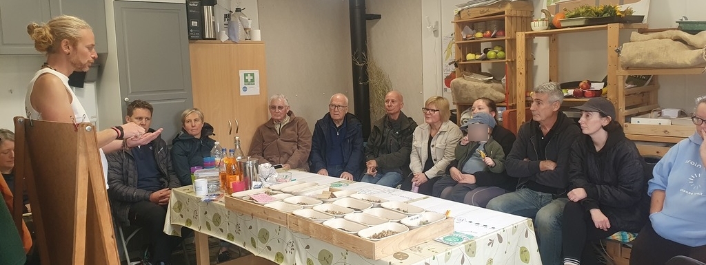 10 adults and a child sit in a line around a table full of boxes of seeds, while a man in a white shirt with a ponytail tells them about the Fife Seed Library