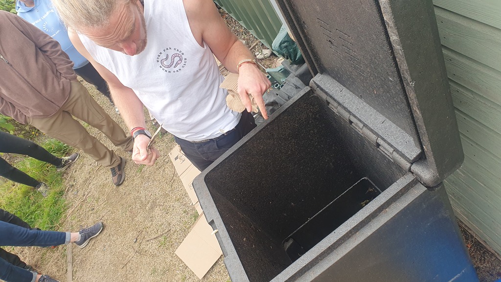 A man in a white shirt with a worm picture points into an open hot compost bin