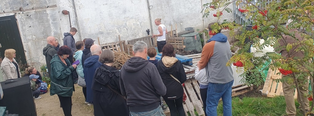 A group of people surround several compost bays in a garden while a man in a white shirt and a ponytail explains them
