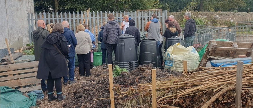 A group of about 17 people stand in a garden while a man in a white shirt explains compost to them. In the foreground are stacked compost bins, a manure pile, and a dead hedge.