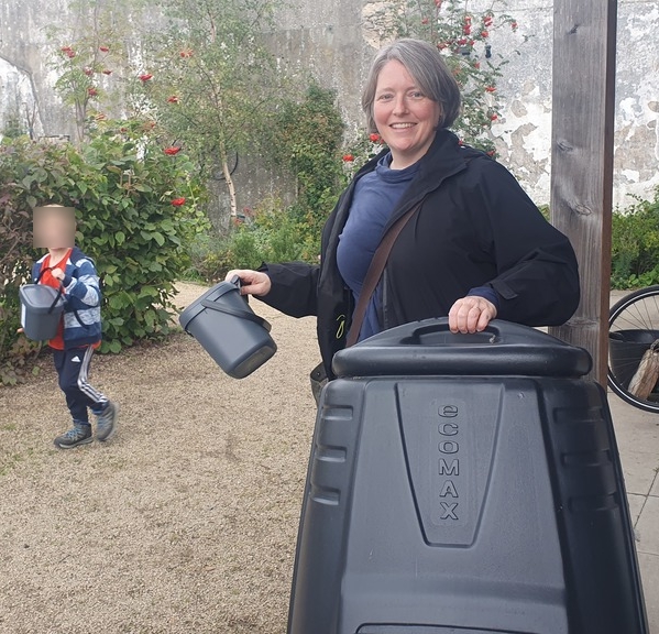 A white woman with grey hair is smiling and holding a large compost bin and a small food waste caddy, on a gravel path with trees in the background. Nearby a small child also carries a food waste caddy.