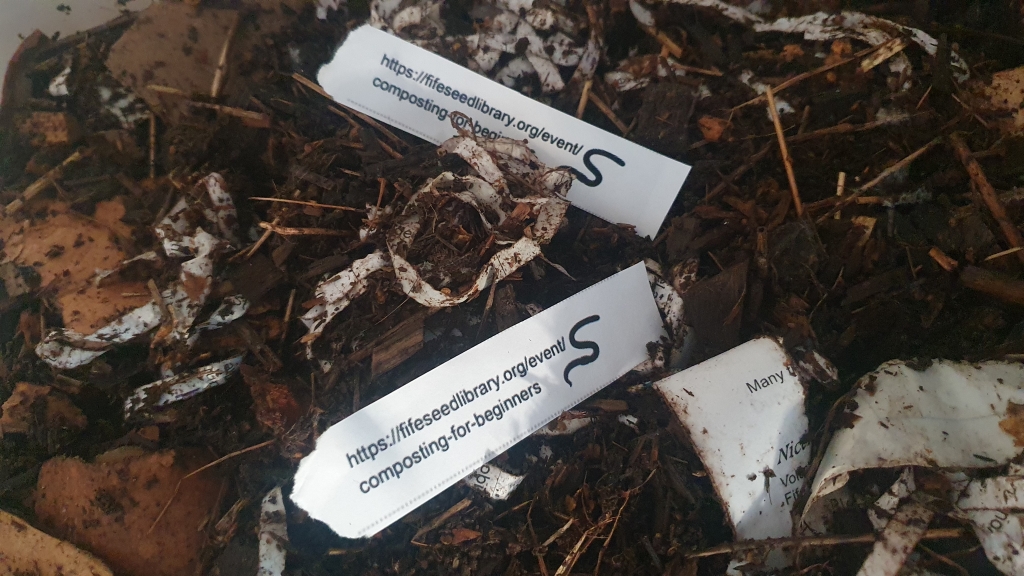 A close up of worm bedding with shredded paper and soil, and two slips of paper with the URL for the composting workshop on