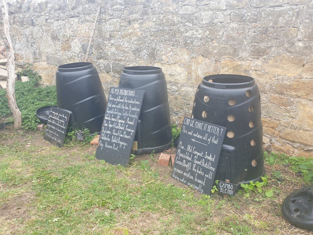 Three black compost bins on grass with handwritten signs leaning against them