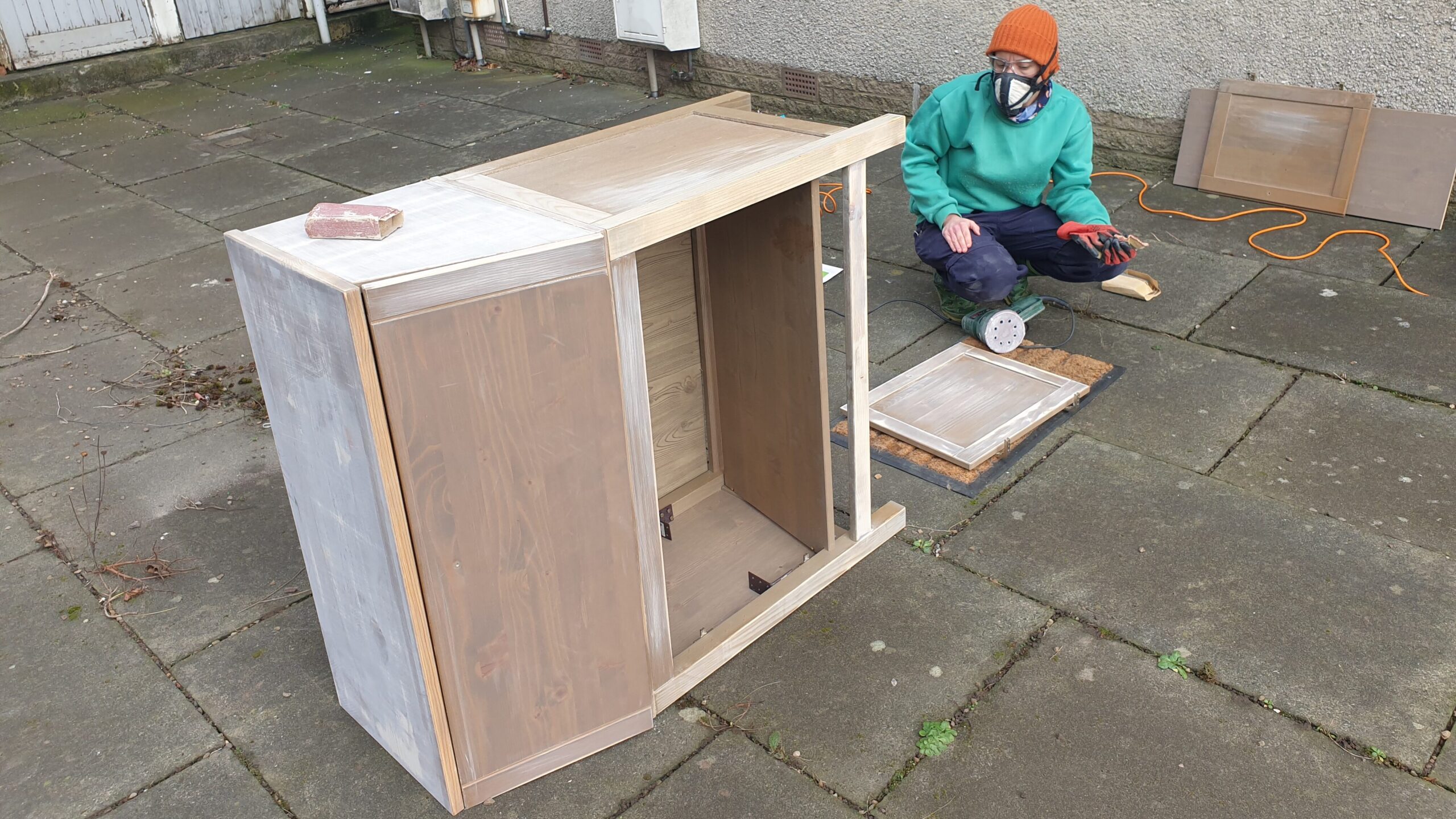 A person with a face mask sanding a wooden cabinet.