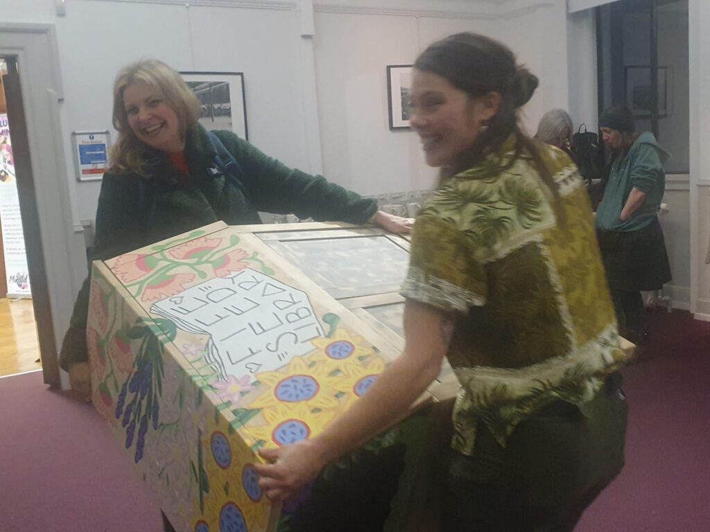 Two people carrying the seed library cabinet on its side, with big wide smiles