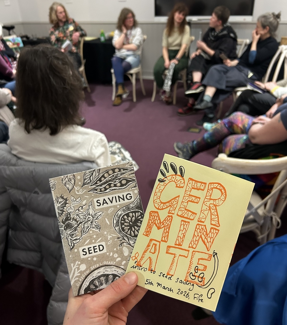 A hand holding two seed zines in front of a group of people sitting in a circle